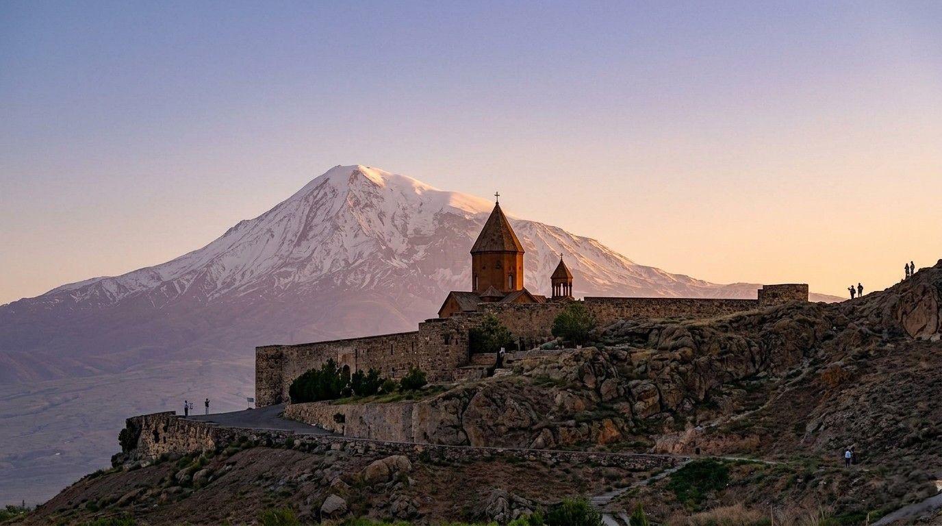 A panoramic landscape photograph taken at sunset, showing the historic Armenian Khor Virap monastery complex perched on a rocky hill, with the massive, snow-covered Mount Ararat looming majestically in the background against a clear, pastel sky.